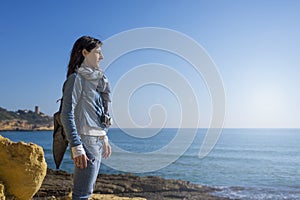 Side view of a daydreaming young woman wearing casual clothes standing on seashore while looking away to the horizon in a bright