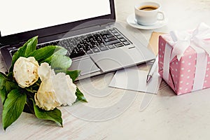 Side view of a deck with computer, bouquet of peonies flowers, cup of coffee, empty card and pink dotted gift box. White rustic wo