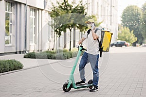 Side view of delivery man in face mask using smartphone and riding push scooter in the city.