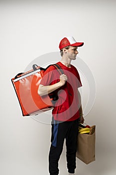 Side view on delivery man in red uniform with thermal backpack and a craft paper bag with groceries. Takeaway food