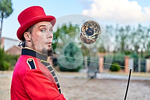 Side view of eccentric bearded man in red costume and hat juggling diabolo and looking at camera