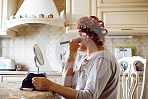 Side view of elderly woman in pink hair-curlers sitting at table, looking at mirror, putting powder rouge on skin.