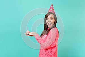 Side view of excited woman in knitted pink sweater, birthday hat keeping mouth open, looking surprised hold cake with