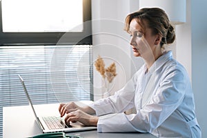 Side view of female doctor physician wearing white coat working typing on laptop computer looking on display screen.