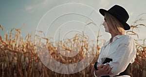 Side view: A female farmer in a dress and hat walks along the fields of ripe corn.