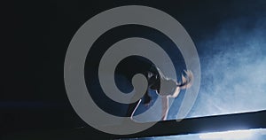 Side view of a female gymnast doing split handstand on balance beam against black background.