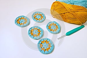 Side view of five round crochet motifs in turosque and yellow colors, a hook and skeins of yarn on smooth white surface