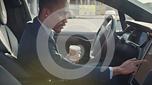Side view of focused african man sitting on driver's seat with cup of coffee and using dashboard for navigation