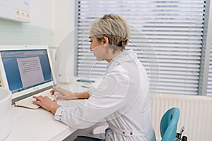 Side view of focused middle-aged woman general practitioner in white coat using laptop computer writing notes at