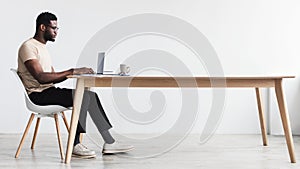 Side view of focused young black man using laptop, sitting at desk, working or studying online from home office