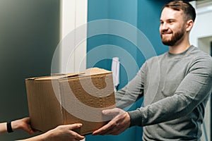 Side view of friendly bearded courier male delivering cardboard box parcel to female customer on doorstep at home.