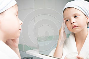 Side view of a girl applying cream on her face while standing in front of a mirror after a shower