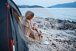 Side view of girl sitting near tent, holding cup of fragrant tea.