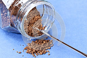 Side view of a glass jar of instant coffee and spoon with some of the granules spilling onto a blue background.