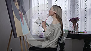 Side view gorgeous female painter making strokes with acrylic paint brush sitting at picture thinking. Talented inspired