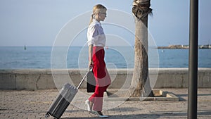 Side view of gorgeous slim confident businesswoman with travel bag walking in slow motion along blue Mediterranean sea