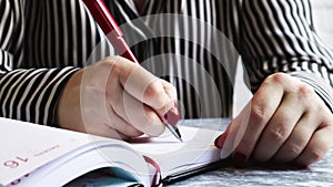 Side view of the hands of a woman with red nails, holding a red pen, writes something on an empty sheet of diary paper lying on a