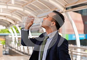 Side view of handsome Asian businessman in suit standing holding mineral water bottle and drinking for health and refresh in heat