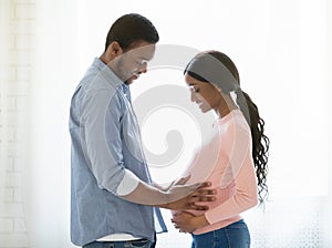 Side view of handsome black man touching his wife`s pregnant belly near window at home