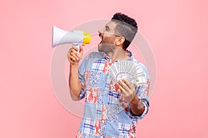 Side view of handsome crazy screaming in megaphone man with beard holding fan of dollars, wearing