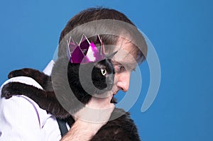 Handsome man in shirt holding on sholder black cat in pink shiny crown on blue background photo