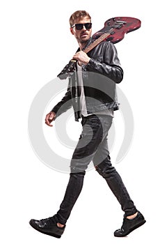 Side view of handsome rocker walking with guitar on shoulder