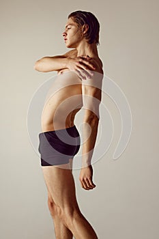 Side view. Handsome shirtless young guy with muscular body standing in underwear against grey studio background. Fitness