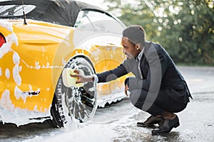 Side view of handsome young African businessman using yellow sponge for cleaning rims outdoors.