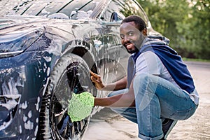 Side view of handsome young African man using green microfiber car wash mitt for cleaning rims of his modern car