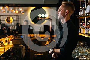 Side view of handsome young barman standing near bar counter waiting for customers orders on blurred background of