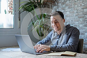 Side view handsome young businessman in eyewear working with computer remotely, sitting at wooden table in office. Pleasant happy