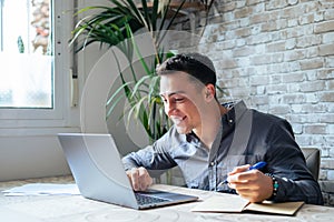 Side view handsome young businessman in eyewear working with computer remotely, sitting at wooden table in office. Pleasant happy