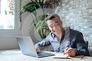 Side view handsome young businessman in eyewear working with computer remotely, sitting at wooden table in office. Pleasant happy