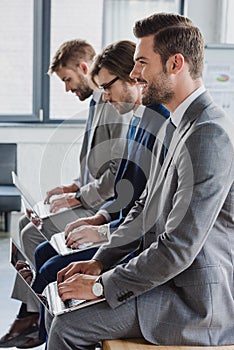 side view of handsome young businessmen in suits sitting and using laptops