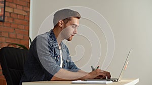 Side view of handsome young man sitting at desk, opening laptop and starts typing on keyboard of computer.