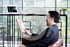 Side view of a happy Asian businessman smiling and reading the newspaper in the morning while sitting on the sofa in the office