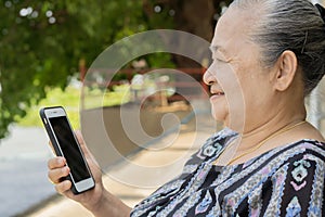 Side view of happy Asian elder senior grey haired woman sitting outdoor and holding mobile phone