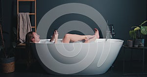 Side view happy beautiful young Caucasian woman relaxing in modern glamour bath, using smart phone shopping online app.