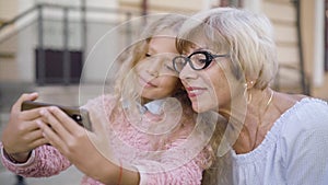 Side view of happy carefree senior woman and pretty little girl making selfie photo on smartphone. Portrait of joyful