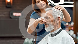 Side view of happy handsome bearded man getting haircut at barbershop. Young barber girl working with hair clipper