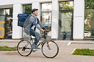Side view of happy handsome young delivery man with thermo backpack riding bicycle in city street on blurred background