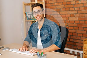 Side view of happy handsome young freelance designer male in stylish glasses working on desktop computer sitting at desk