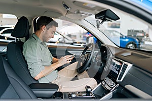 Side view of happy handsome young man sitting behind wheel of luxury car and using mobile phone for surfing internet