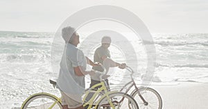 Side view of happy hispanic senior couple walking with bikes on beach at sunset