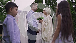 Side view of happy Middle Eastern woman and Caucasian man talking on wedding day with blurred children chatting at front