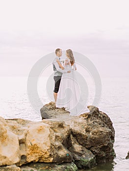 The side view of the happy newlyweds standing on the end of the cliff among the sea.