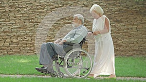 Side view of happy senior wife rolling disabled husband in wheelchair in sunny park. Wide shot of relaxed old Caucasian