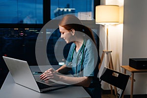 Side view of happy smiling redhead young female doctor in blue green medical uniform working typing on laptop computer.