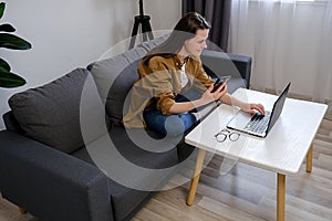 Side view of happy young caucasian woman 20s using mobile phone while sitting on cozy couch at home with laptop computer. Internet