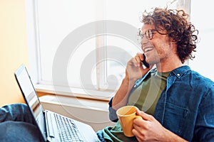 Side view of happy young man sitting in the office talking on mobile phone while working on a laptop.